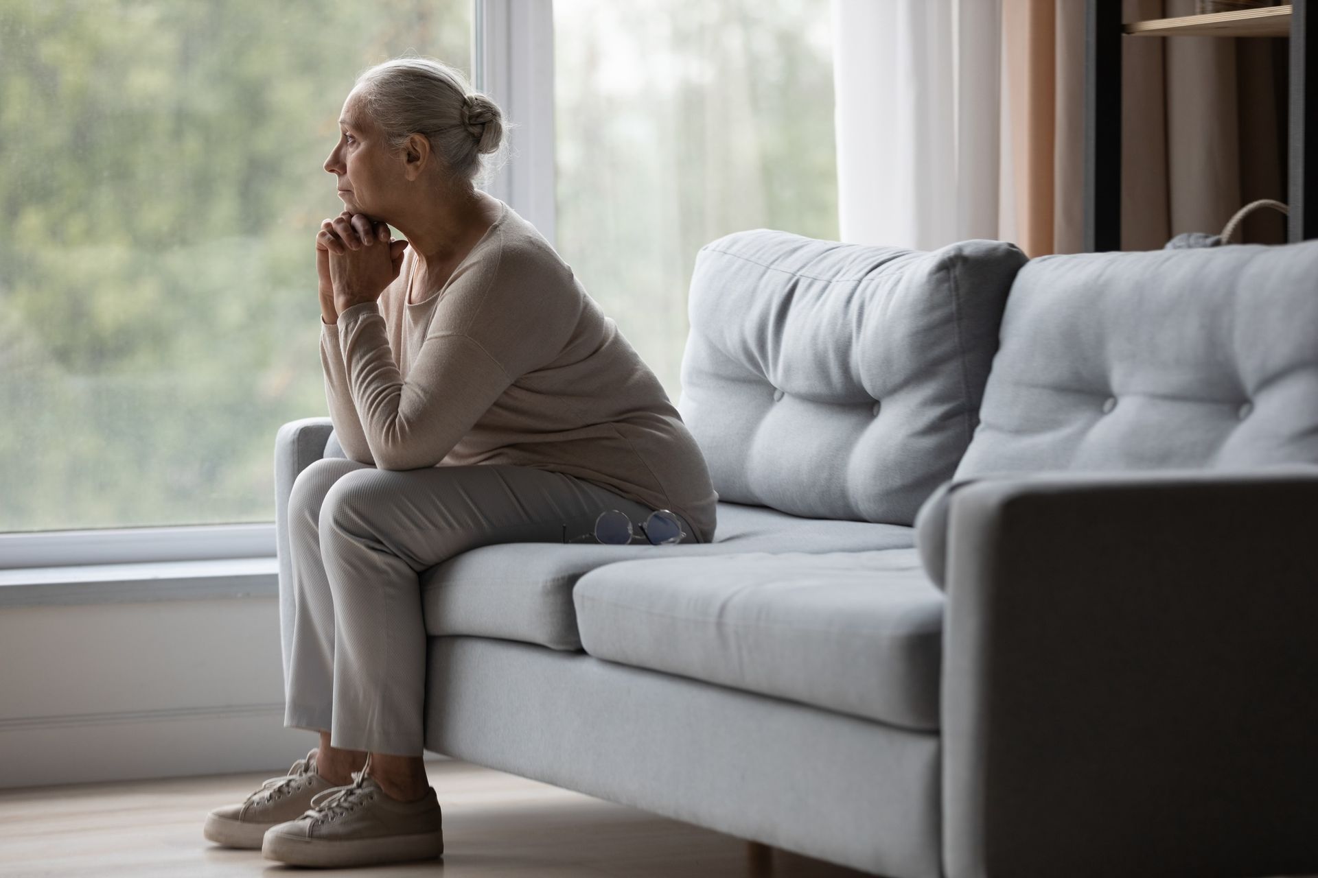 An elderly woman is sitting on a couch looking out a window.