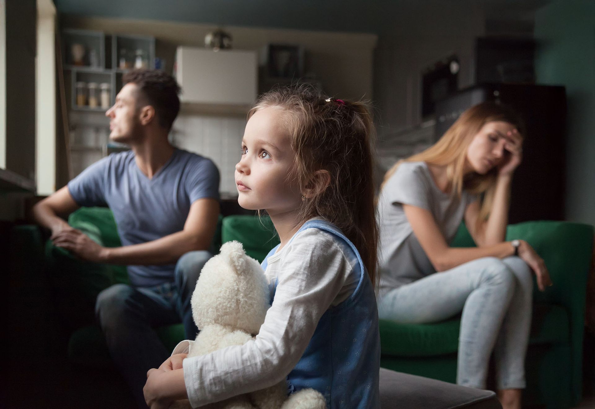 Child hugging a teddy bear, looking up, with parents on a couch in the background, seeming unhappy.