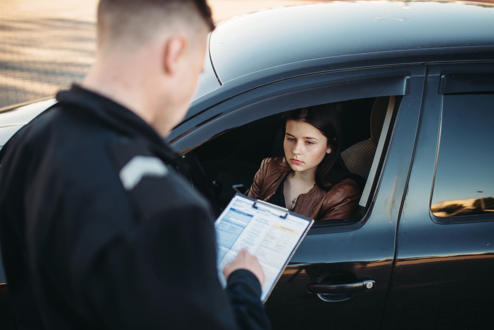 Police officer issuing a traffic ticket to a young woman in a car at dusk.