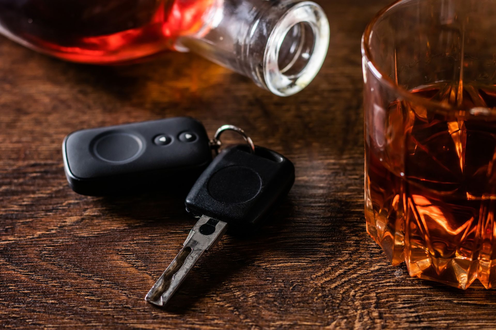 Car keys and remote next to a glass of amber liquor and a bottle, on a wooden surface.