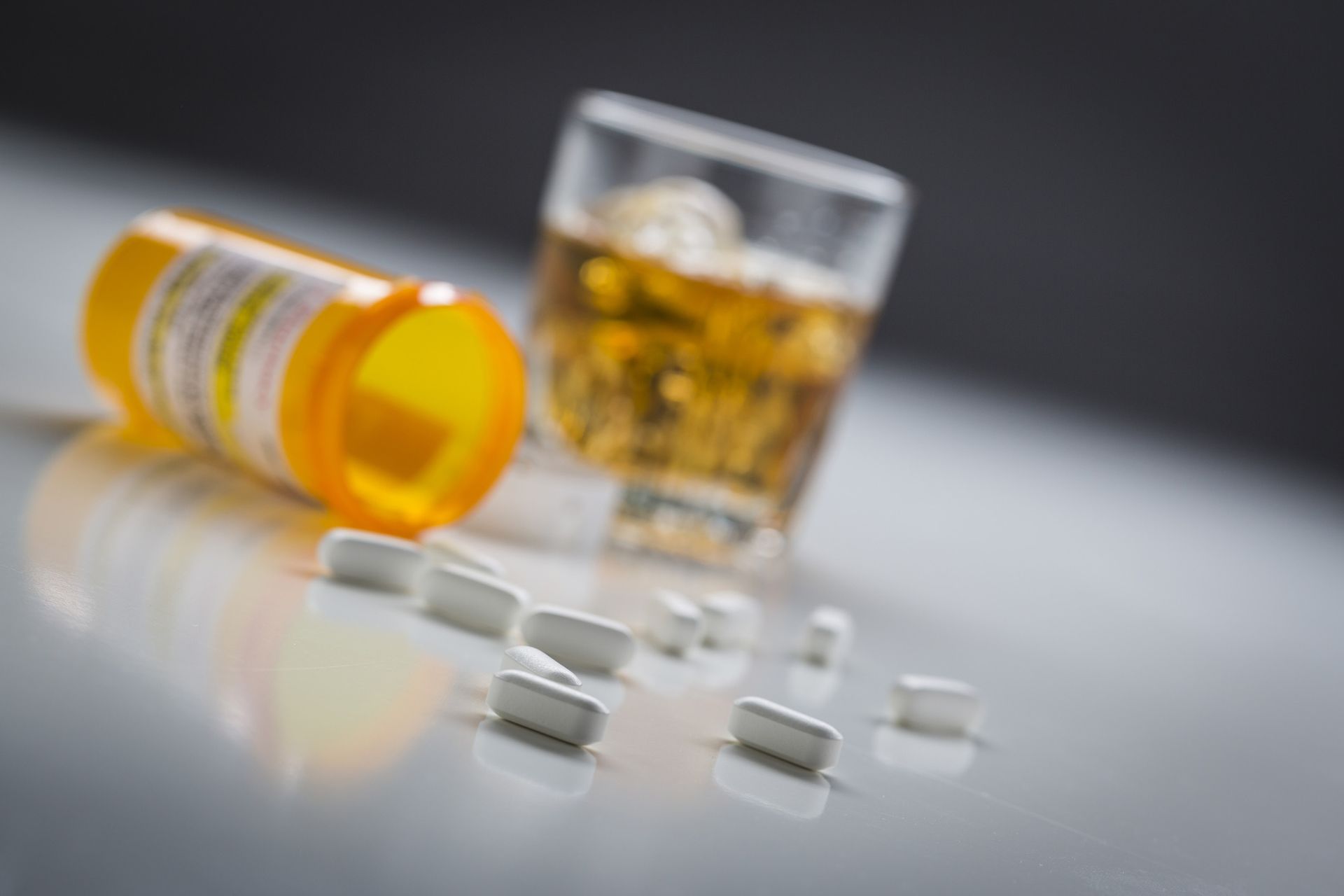 Yellow pill bottle and white pills spilled on a surface next to a glass of amber liquid.
