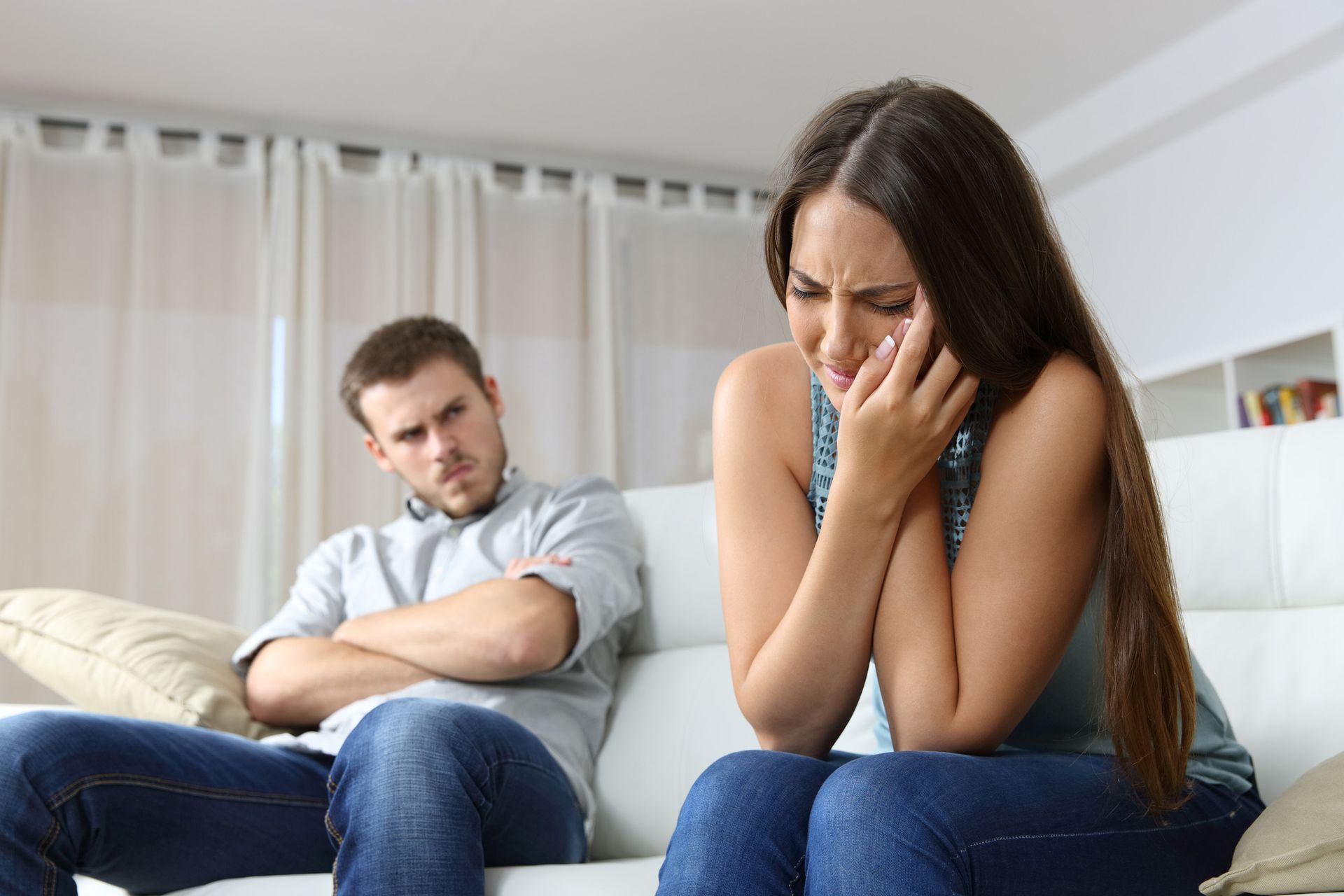 Woman crying on a couch as a man glares at her with arms crossed in a living room.