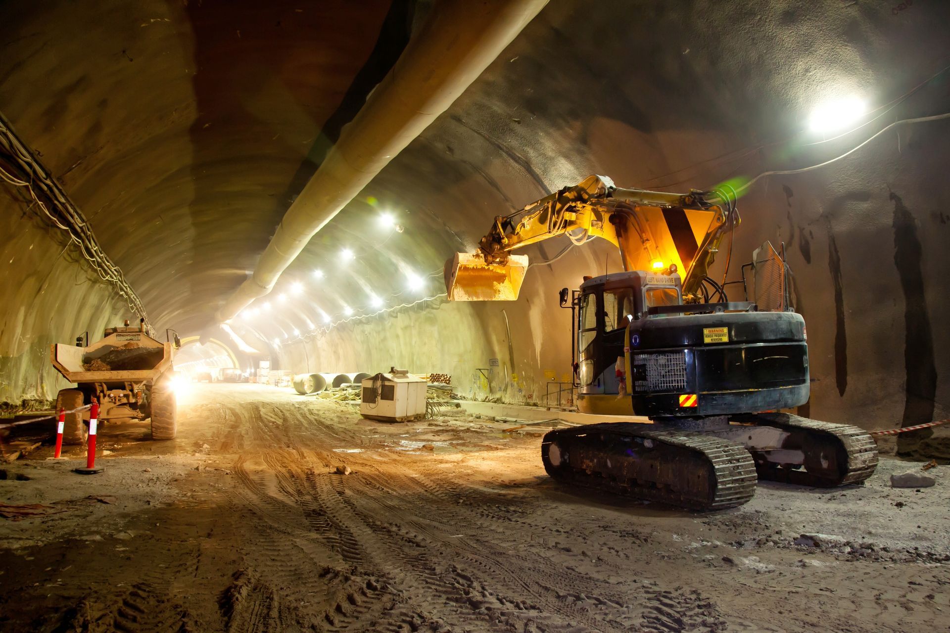 Construction of a tunnel: Excavator and truck inside a dark tunnel, lit by work lights.