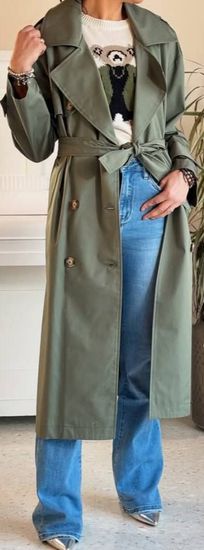 Una persona che indossa un trench verde oliva, jeans azzurri e tacchi è in piedi all'interno.