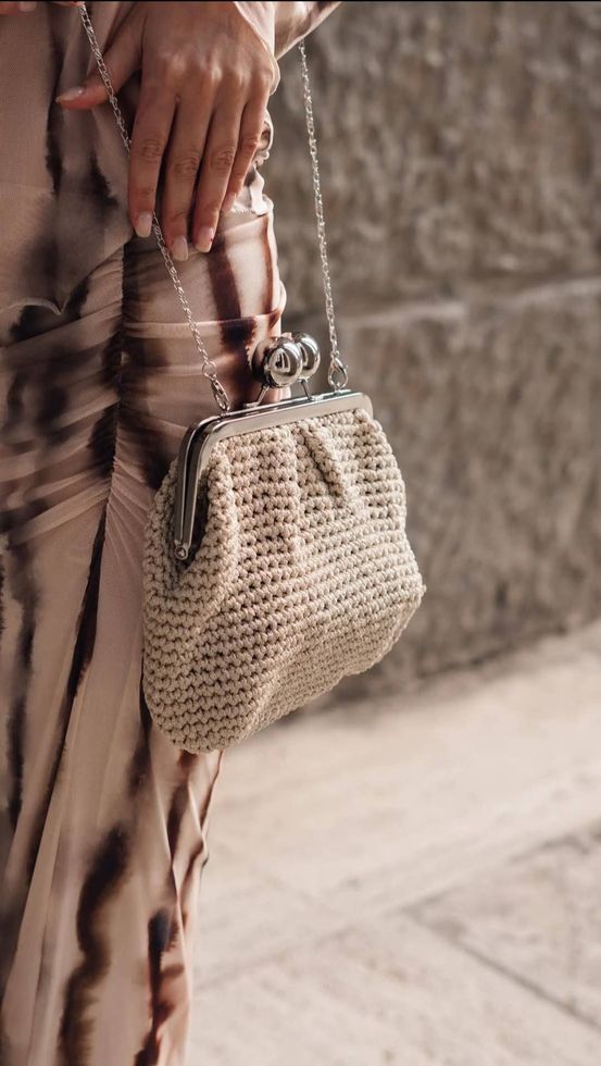 Donna che tiene in mano una borsa beige intrecciata con tracolla a catena argentata e chiusura, e indossa un abito tie-dye.