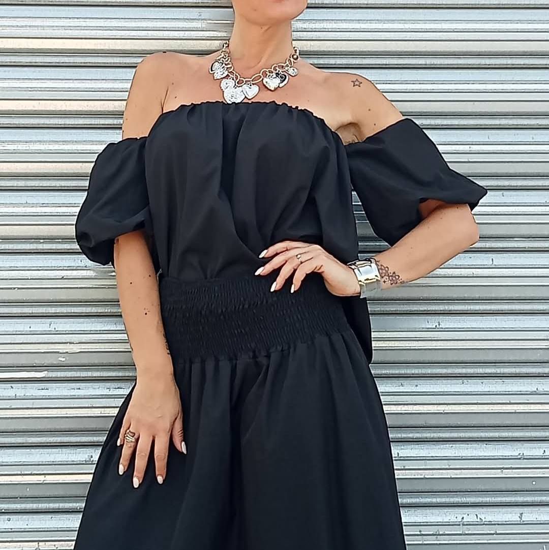 Donna con abito nero a spalle scoperte, collana e braccialetto, in posa davanti a una persiana argentata.