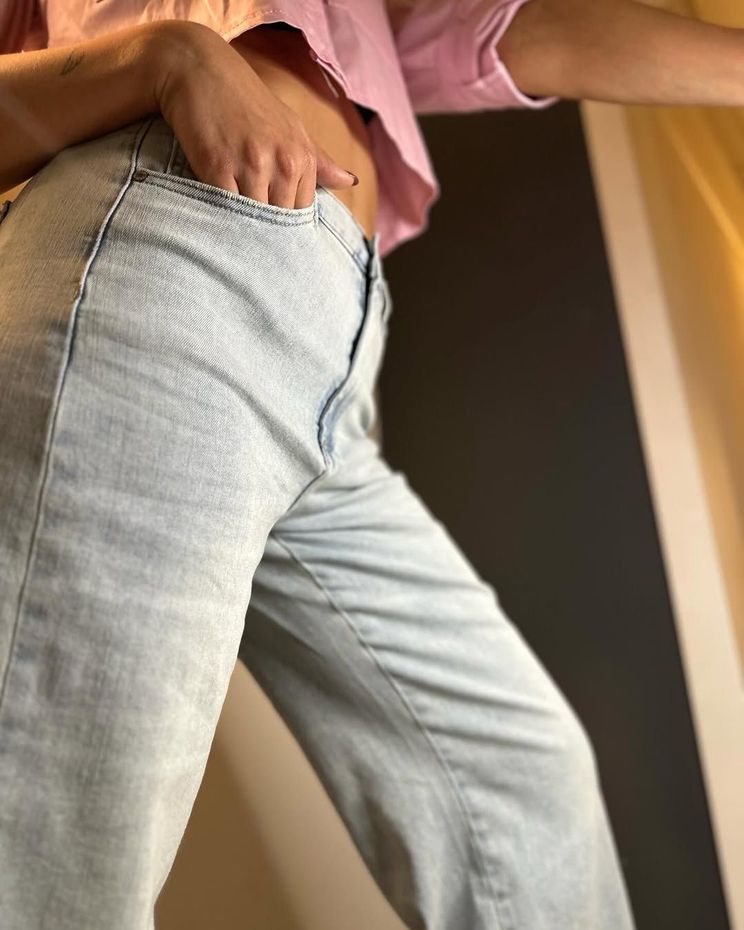 Donna in piedi sulla soglia di una porta, con una mano in tasca, che indossa un top corto rosa chiaro e jeans azzurri.