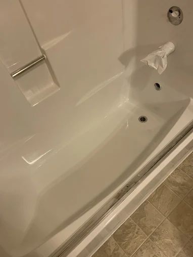 Bathtub with beige tile floor, built-in grab bar, and a broken faucet handle near the drain.