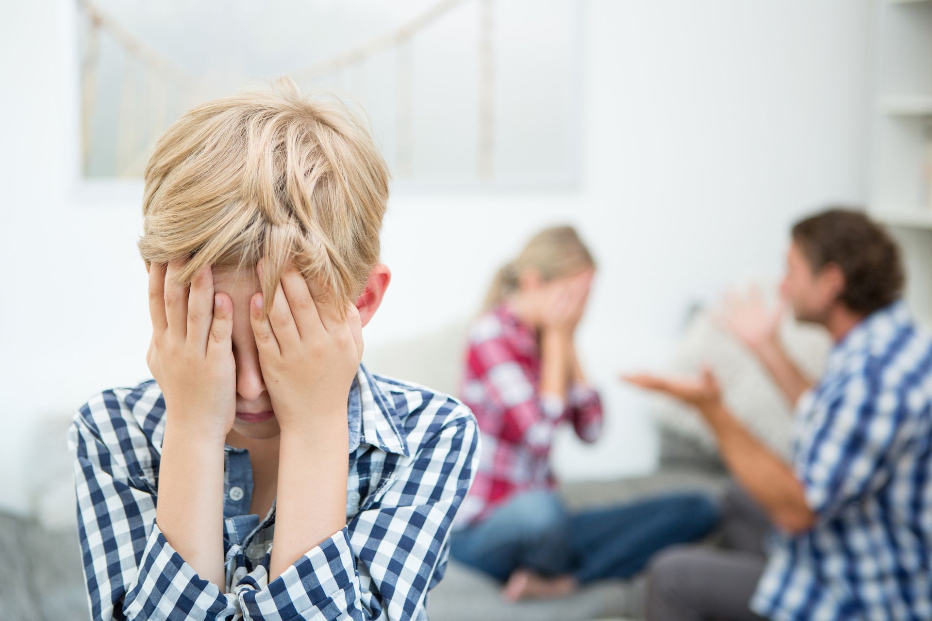 A boy is covering his face while his parents argue in the background.