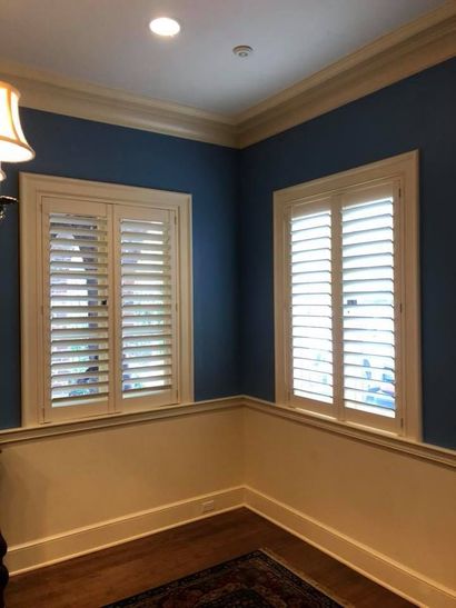 Two white shuttered windows in a corner room with blue walls above cream-colored wainscoting.