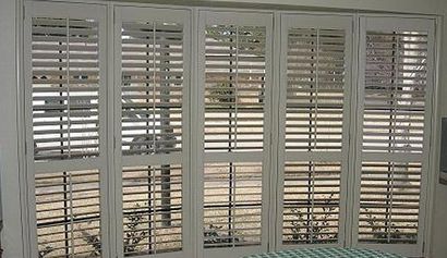 White shuttered windows, partially open, overlooking a garden. The shutters are divided into upper and lower sections.