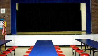 A school cafeteria with a stage at the back, blue tables, red stools, and an American flag.