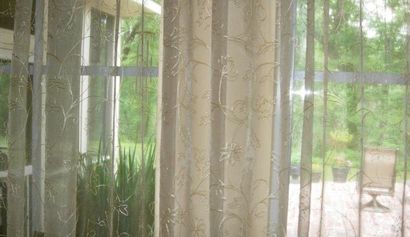 Sheer beige curtains hang in front of a glass door or window with a view of green foliage and a patio.
