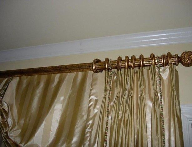 Gold curtain rod with rings holding up pleated, striped gold curtains against a cream-colored wall.