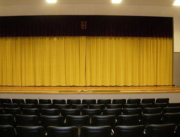 Stage in an auditorium with closed yellow curtains, maroon top curtain, and rows of black chairs.