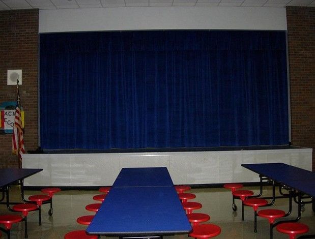 A school stage with a closed blue curtain, tables, and red stools in a cafeteria setting. Brick walls are visible.