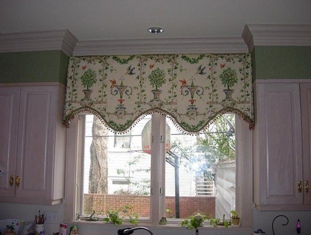 A kitchen window with a decorative valance featuring floral and bird patterns. The valance is cream-colored with green and red accents, and the room has light pink cabinets.