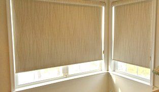 Two closed beige roller blinds in a corner window, filtering sunlight. White window frames and a light beige wall are visible.
