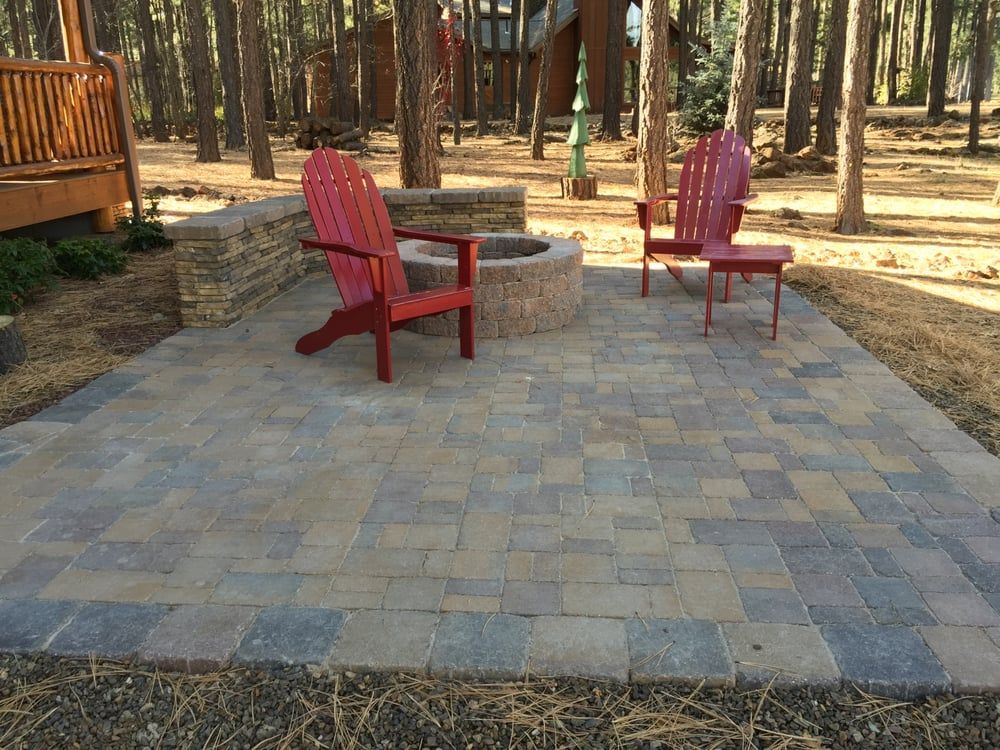 Brick patio with two red Adirondack chairs near a fire pit, set in a wooded area.