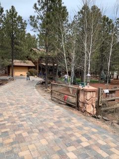 Brick paved driveway leads to a house with wooden fence and trees.