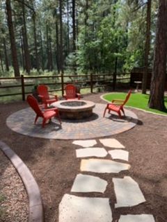 Circular stone fire pit area with red chairs, flagstone path, and forest backdrop.