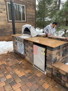 Outdoor kitchen with granite countertop and brick facade, includes grill, refrigerator, and pizza oven. Man working.