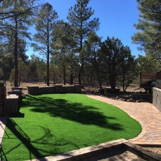 Green artificial turf backyard with stone wall and walkway. Trees and blue sky in the background.