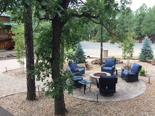 Patio with fire pit, blue chairs, surrounded by trees, gravel, and a paved walkway.