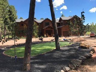 Log home with landscaping in progress; trees in the foreground, green lawn, gravel border, blue sky.