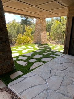Stone patio with stepping stones on artificial turf under a covered structure with stone pillars, overlooking a forest.
