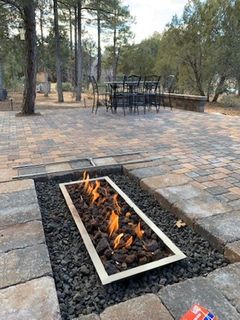 Outdoor fire pit with flames, set in dark lava rocks, surrounded by a stone patio. A table and chairs are in the background.