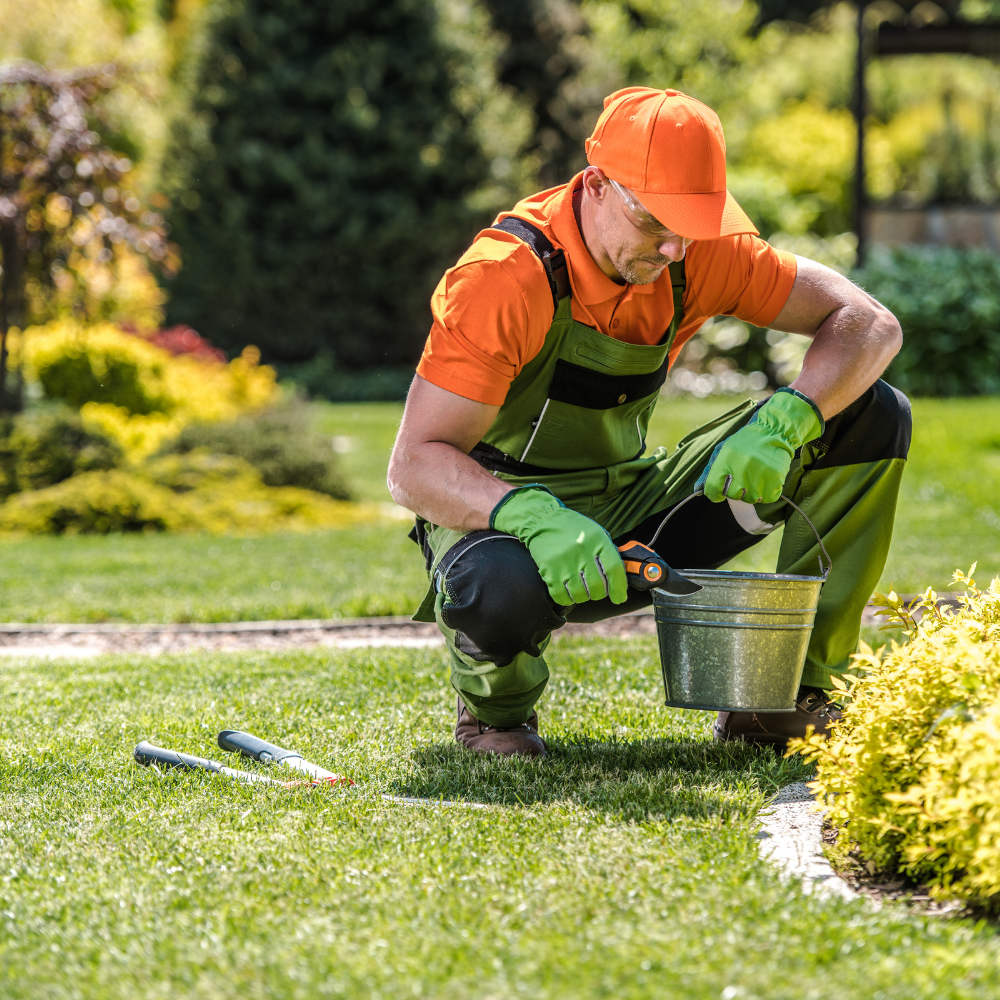 Gardener kneeling in a yard, trimming plants, wearing orange shirt, green overalls and gloves, and an orange hat.