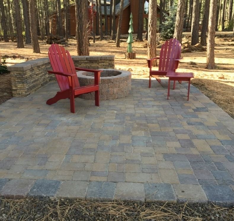 Red Adirondack chairs surround a fire pit on a paver patio in a wooded area.