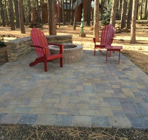 Red Adirondack chairs surround a fire pit on a paver patio in a wooded area.
