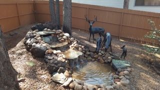 Water feature with a small waterfall, pond, and deer sculptures, in a yard with a wooden fence.