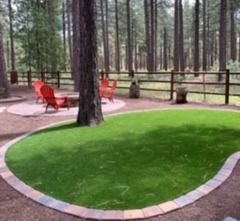 Circular patch of artificial turf bordered by bricks, with red chairs and a fire pit in a wooded area.