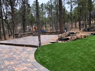 Brick patio and green lawn in a wooded setting; small waterfall feature, two people in the background.