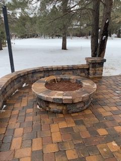 Brick patio with a circular fire pit, snowy background, trees.