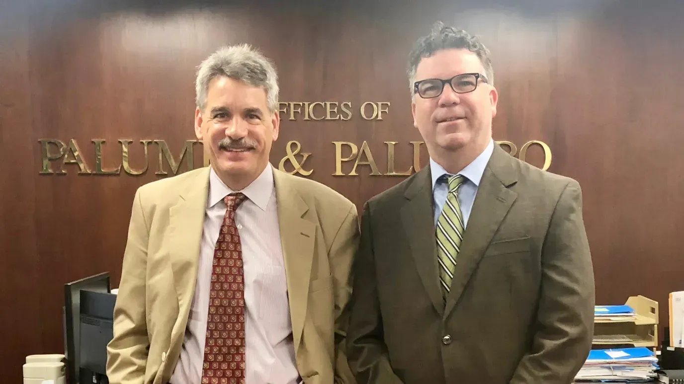 Two men in suits stand in front of a sign that reads