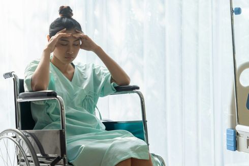Woman in hospital gown, sitting in a wheelchair, holding her head in apparent distress near a window.
