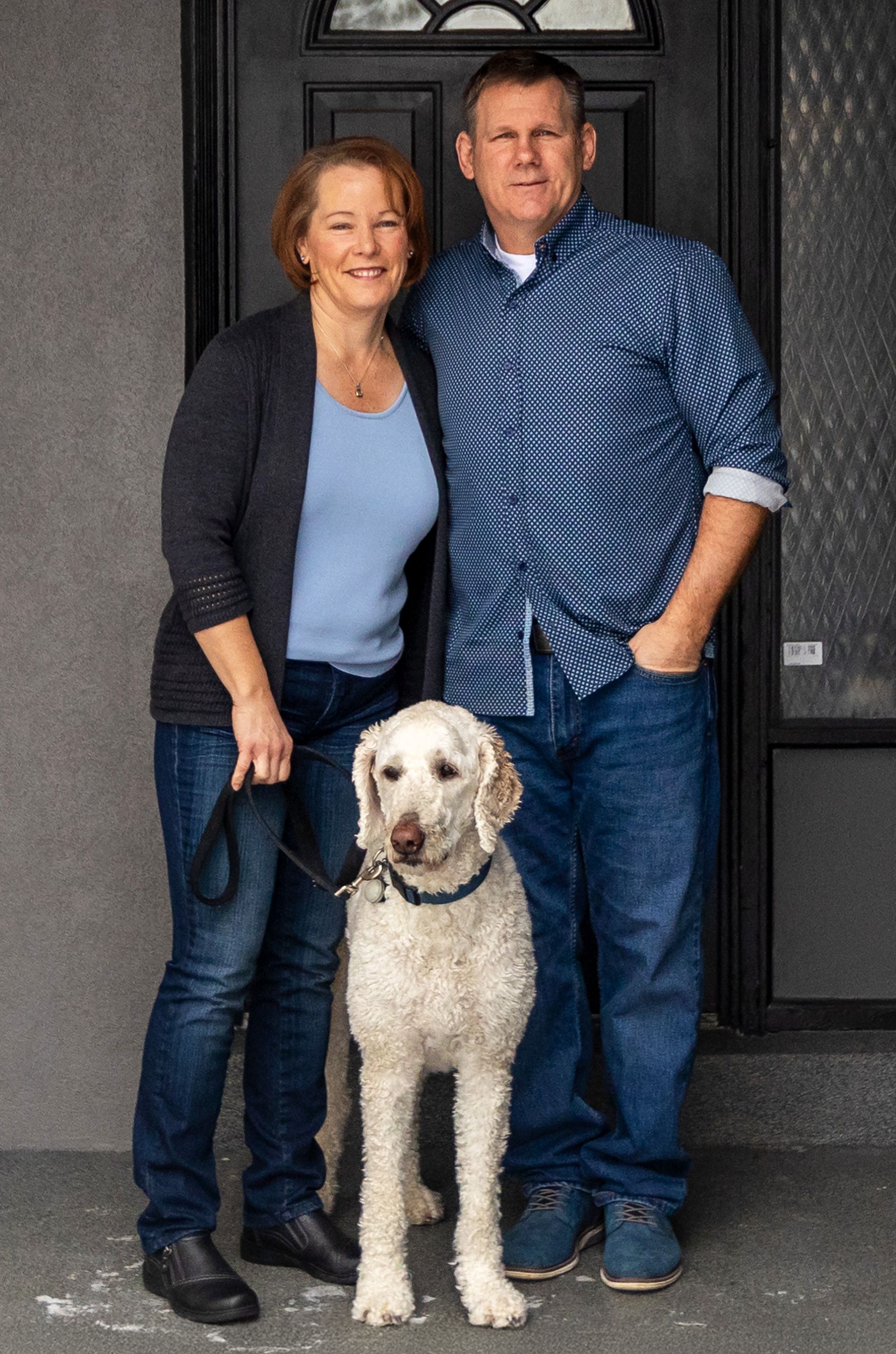 Woman and man stand with a fluffy white dog in front of a dark doorway. They are smiling.