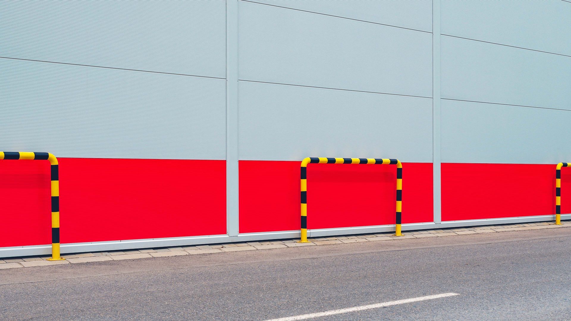 Red and gray industrial building with yellow and black safety barriers along a paved road.