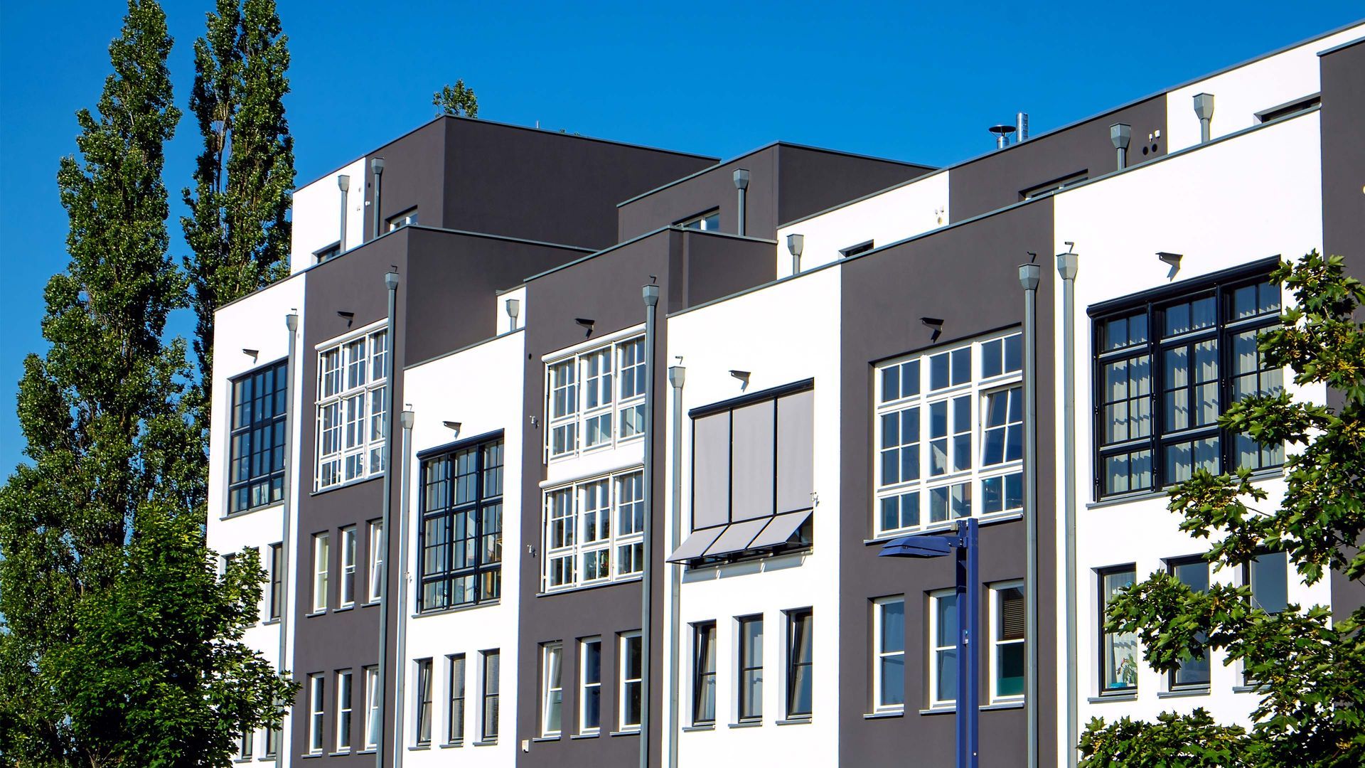 Modern multi-story building with white and gray facades, rows of windows, and clear blue sky.