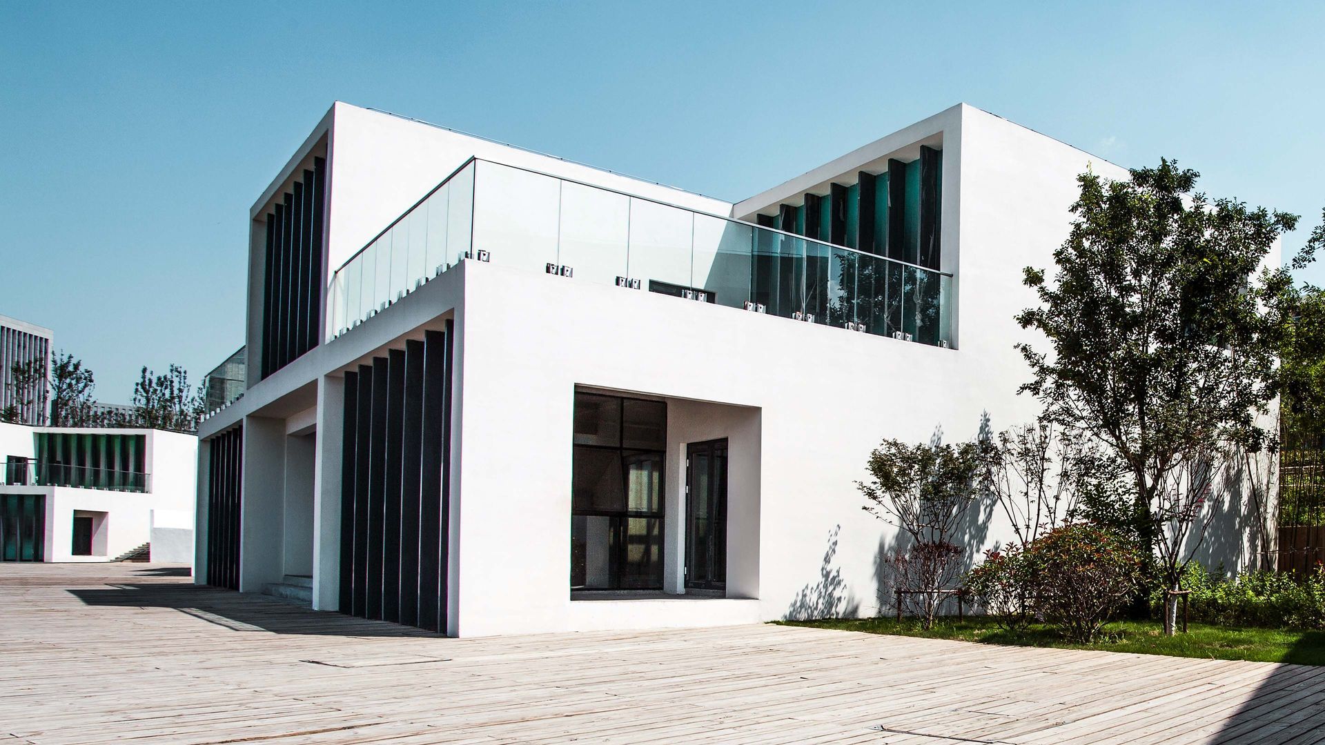 White modern building with large windows and a glass balcony, set on a stone paved area with greenery.