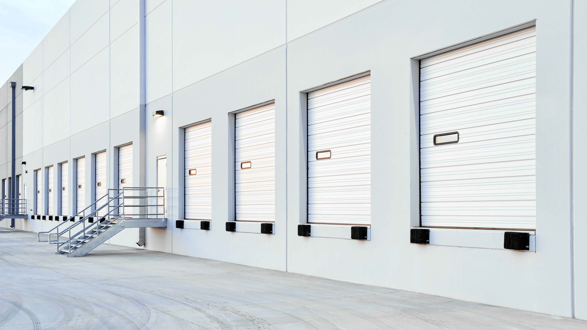 Exterior of a warehouse with several loading dock doors; white building, light gray pavement, steel staircase.