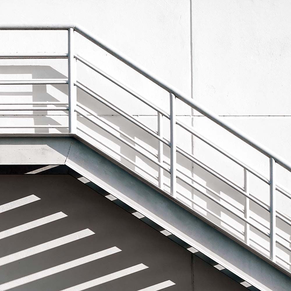 White metal staircase with angled railings against a white wall; shadows cast on the surface below.