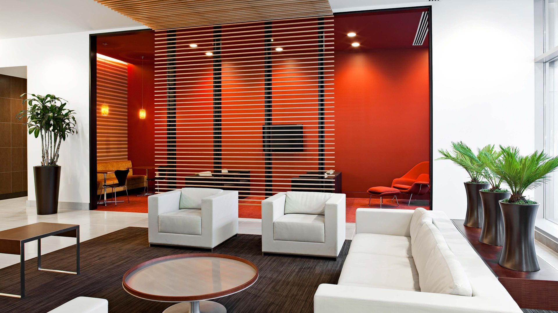 Modern office waiting area with white furniture, red accents, and potted plants.
