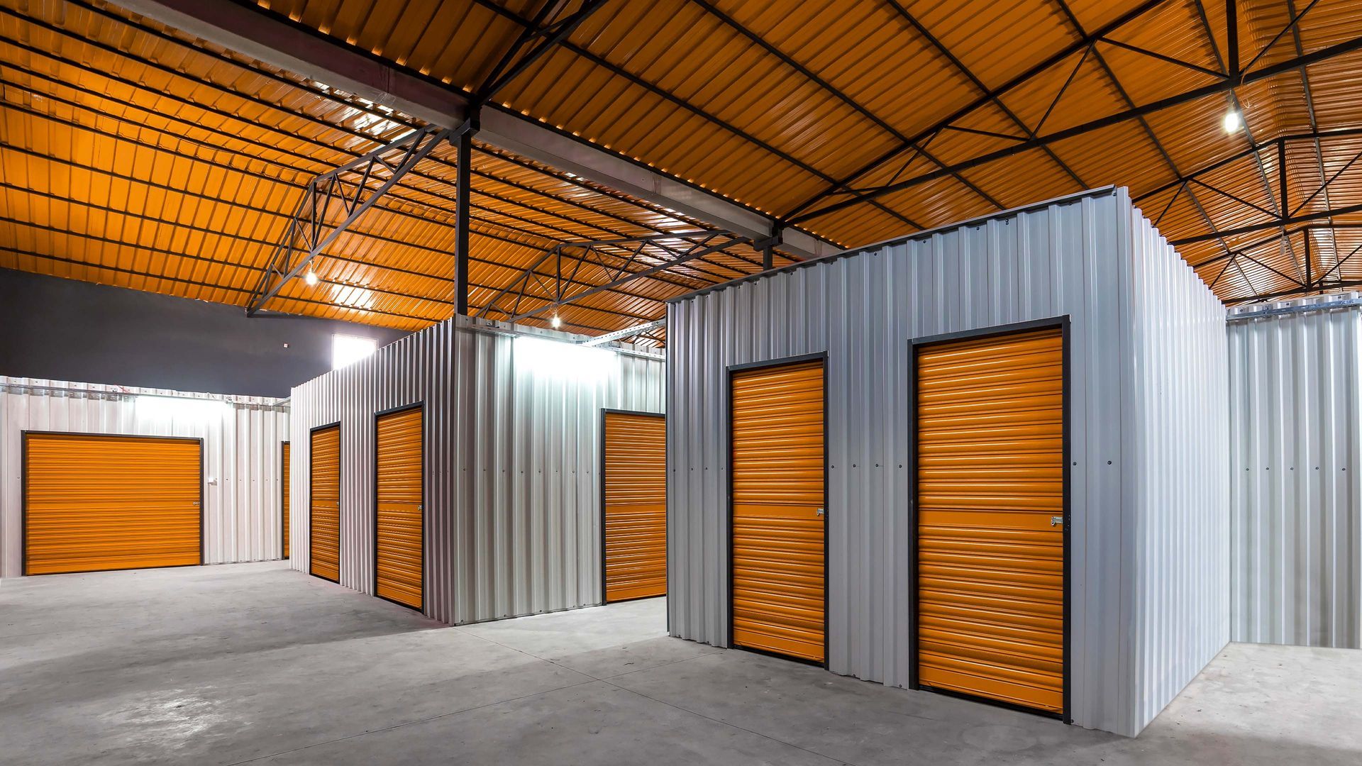 Storage units with orange doors and silver metal siding in a large warehouse setting.