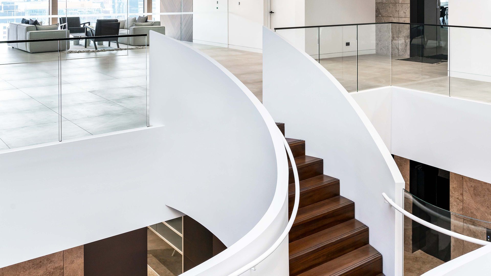 Curving white walls and wooden stairs in modern interior; glass railing; lounge seating visible on the upper level.