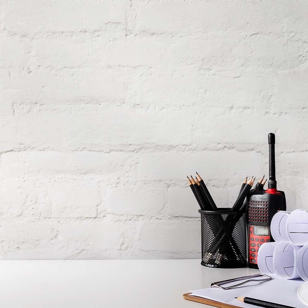White desk with pencils, walkie-talkie, blueprints, and clipboard against white brick wall.